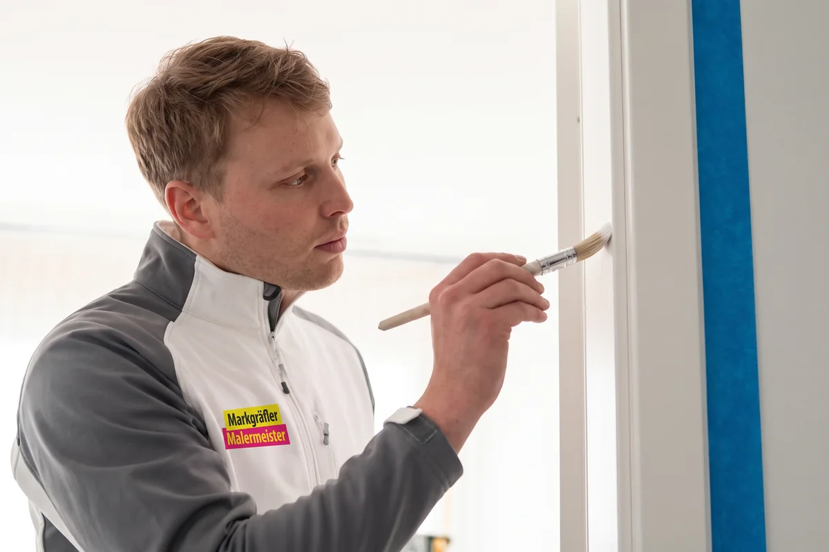 Ein Maler in Schutzkleidung führt mit einer Lackierpistole die Sprühlackierung eines Türrahmens durch