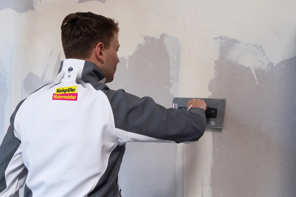 Bärtiger Handwerker in Arbeitskleidung glättet eine Wand mit Gips und Spachtel im unfertigen Zimmer