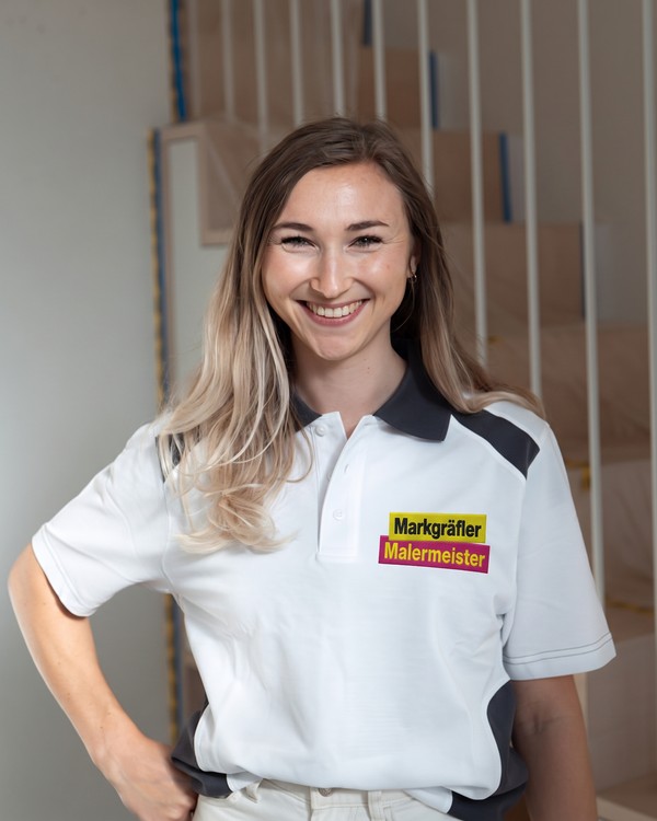 Leona, Planung und Raumdesign, lächelnd im Markgräfler Malermeisterbetrieb Polo-Shirt vor einem unfertigen Holztreppenhaus