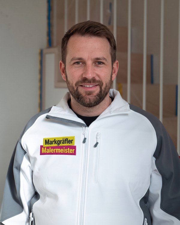 Volker, Teamleiter und Maler, bärtig in Arbeitsjacke des Markgräfler Malermeisterbetriebs vor hellen Wandelementen