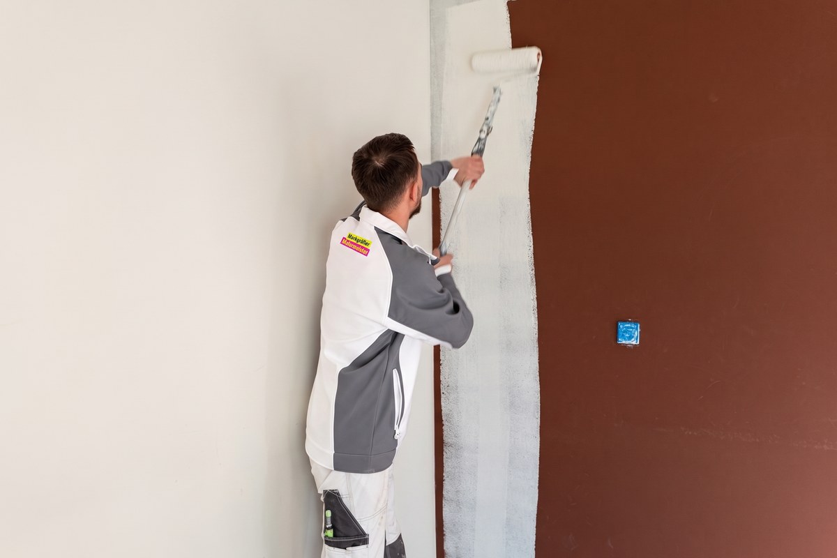 Zwei Maler in Markgräfler Malermeisterbetrieb Arbeitskleidung streichen die Wände eines Raumes, Fenster und Boden sind abgeklebt